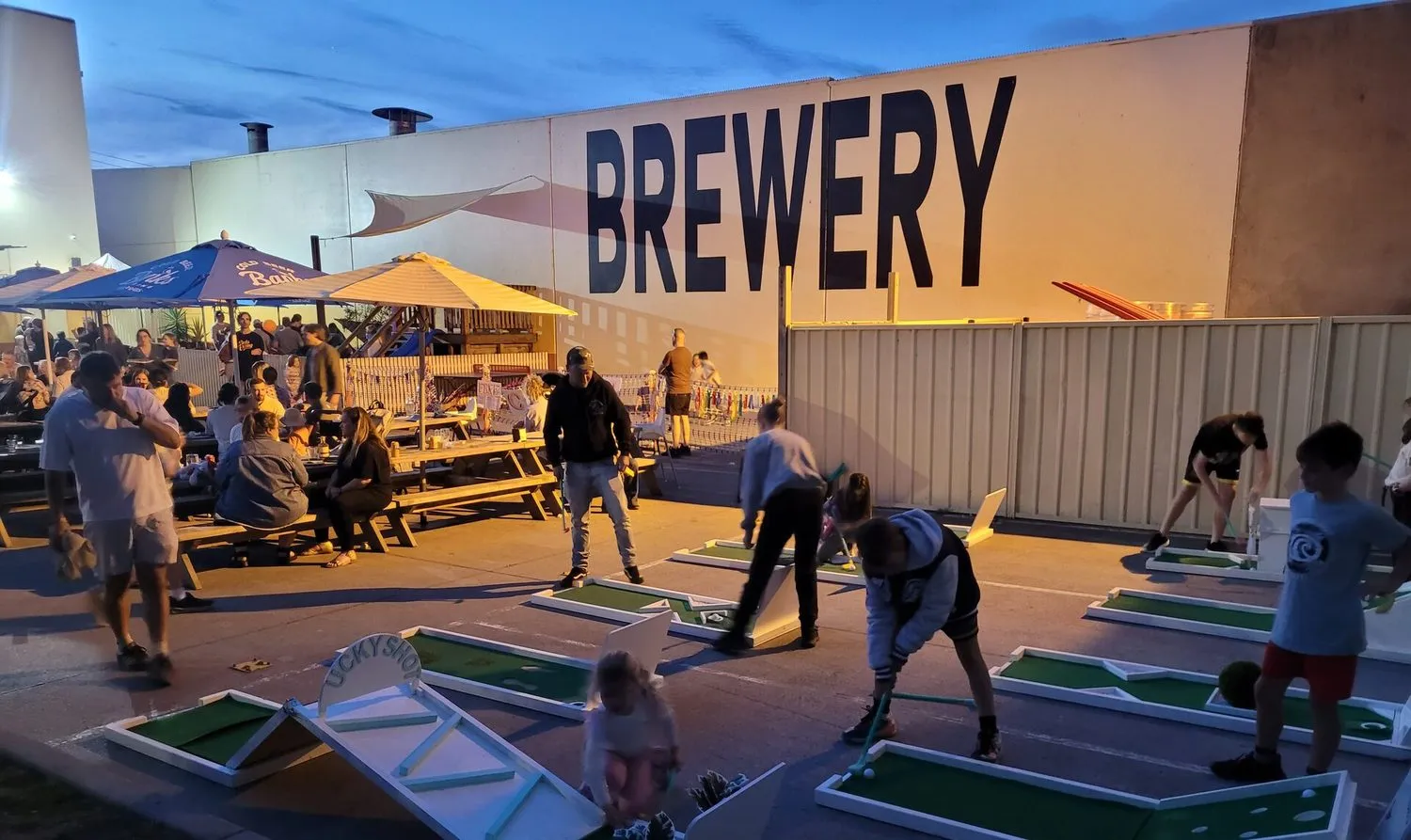 Lucky Shot mini golf course at a Melbourne brewery community evening event, twilight sky and warm picnic-table lighting